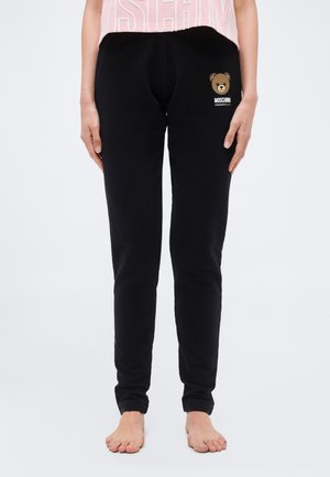 Osoba nosiaca čierne tepláky s logom medvedíka Moschino na stehne, v kombinácii s krátkym ružovým topom, stojaca bosá na bielom pozadí.