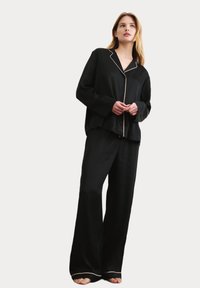Svart satinpyjamas-set med noterat krage, långa ärmar och ljusfärgad pipning; löst sittande byxor med elastisk midja.