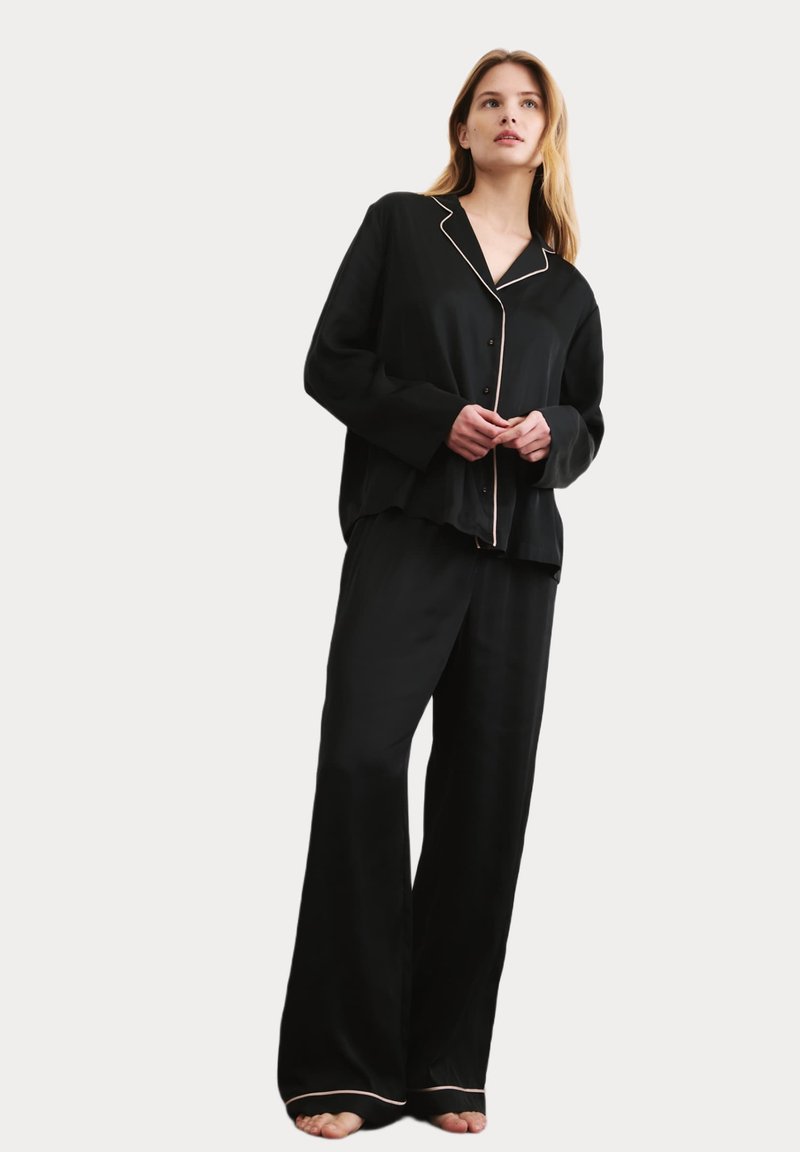 Svart satinpyjamas-set med noterat krage, långa ärmar och ljusfärgad pipning; löst sittande byxor med elastisk midja.