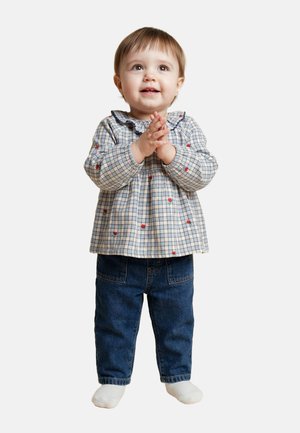 Tout-petit debout, les mains jointes, portant un chemisier à carreaux avec des détails floraux, un jean bleu et des chaussettes blanches sur fond blanc.