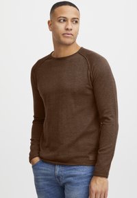 Brun langermet skjorte med ribbet tekstur og raglanermer. Kledd med blå jeans, som har en liten merkeetikett nederst.