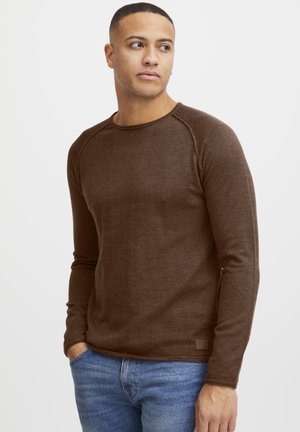Brun langermet skjorte med ribbet tekstur og raglanermer. Kledd med blå jeans, som har en liten merkeetikett nederst.