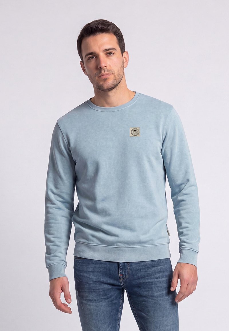 Homme portant un sweat-shirt bleu clair à manches longues et un jean bleu, debout devant un fond clair uni.