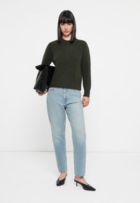 Grønn strikkegenser, lys blå rette jeans, svarte hæler, holder en svart skinnveske. Minimalistisk stil, enkle linjer og teksturer.
