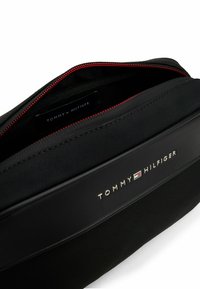 Zwarte cosmeticatas met een textuur op de buitenkant, rode rits en gouden "TOMMY HILFIGER" logo aan de voorkant. Binnenin is het merklabel zichtbaar.