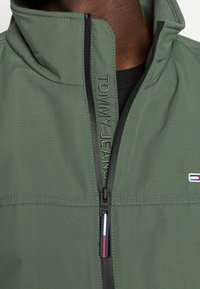 Olivgrön jacka med dragkedja och hög krage, med präglad text "TOMMY JEANS", svart dragkedja och liten logotypmärke med röda, vita och blå accenter.