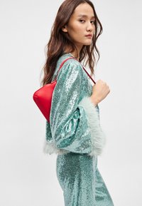 Bolsa de mão triangular vermelha com uma textura suave. O modelo veste uma roupa cintilante em azul-turquesa com detalhes felpudos brancos nas mangas.