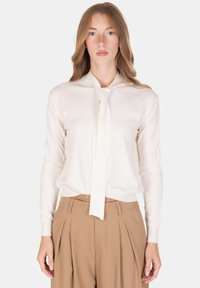 Blusa color crema a maniche lunghe con fiocco al collo, caratterizzata da una texture liscia e una silhouette aderente, abbinata a pantaloni beige a vita alta.