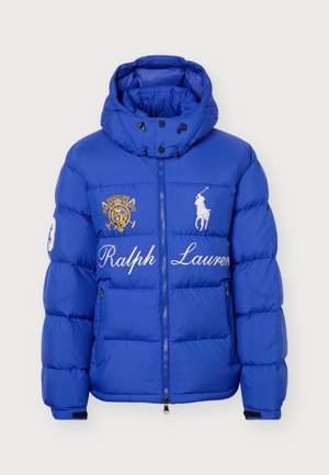 Un mannequin masculin se tient devant un fond uni et clair, vêtu d’une doudoune bleue Ralph Lauren, d’un jean ample et de baskets blanches.