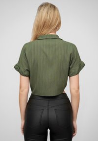 Cămașă verde cu mânecă scurtă și guler, cu dungi verticale subtile și manșete răsucite, asortată cu pantaloni negri ajustați.