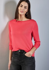 Haut en tricot corail à manches trois-quarts, col rond et poignets côtelés, associé à un jean en denim foncé. Texture lisse avec un minimum de détails.
