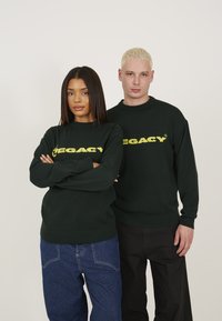 Grüne Strickpullover mit gelbem "LEGACY"-Text auf der Brust, getragen mit Jeans und schwarzen Hosen, beide mit gerippten Bündchen und Säumen.