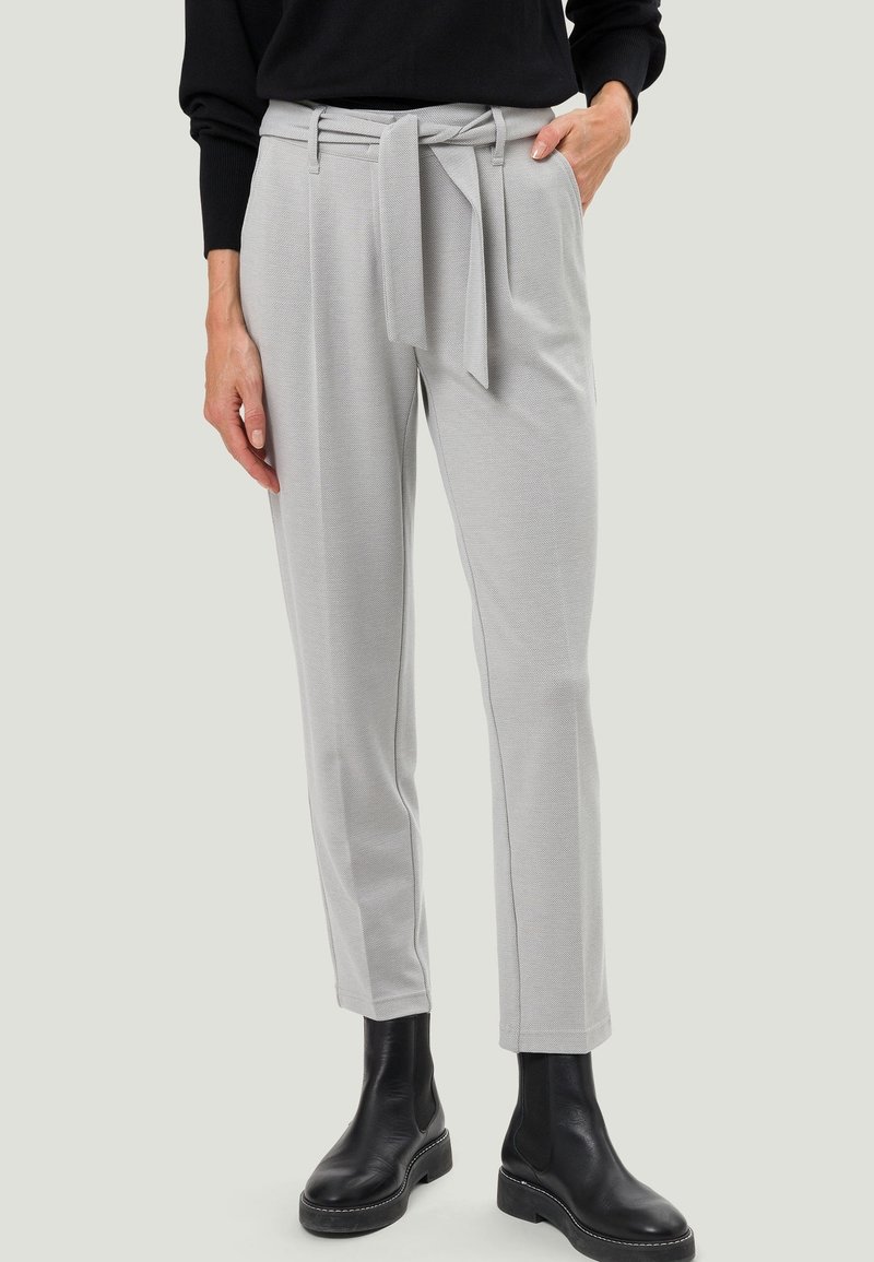 Pantalon gris clair ajusté avec une ceinture en tissu nouée à la taille, porté avec des bottines noires et un haut noir à manches longues.