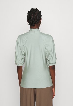 Person mit geflochtenem Haar, die ein hellgrünes Oberteil mit Puffärmeln und braune Hosen trägt, von hinten vor einem einfarbigen Hintergrund gezeigt.