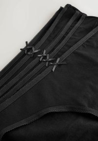 Biancheria intima nera a vita alta con tre pannelli verticali rifiniti in pizzo e quattro piccoli fiocchi di raso su sfondo chiaro.