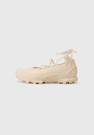 Beige athletic shoe with a smooth, stretch upper, secure lace system, textured sole, and rugged tread for grip. Minimalist design.