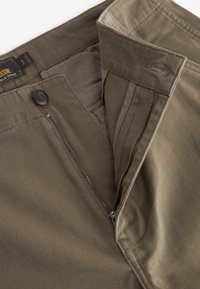 Pantaloni di cotone marroni con zip e chiusura a bottone. Presentano una texture liscia e cuciture pulite lungo la vita e le tasche.