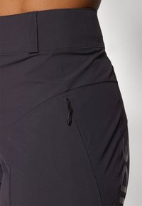 Fekete sport leggings magas derekkal, oldalsó cipzáras zsebbel és sima textúrával, amelyen diszkrét logó található az oldalon.