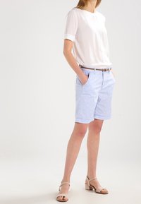 Světle modré šortky s jemným vzorem, spárované s bílou blůzou s krátkým rukávem. Oblečení doplňuje hnědý opasek a béžové sandály.