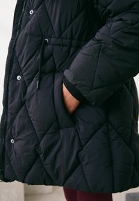 Must tekstitud sulejope tekstuurse pinnaga, millel on hõbedased trukid, reguleeritav kinnituspael ja küljetasku.