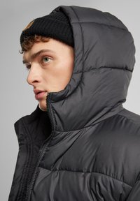 Sort puffjakke med lynlås og høj krave, med et quiltet design. Akkompagneret af en sort strikket hue.