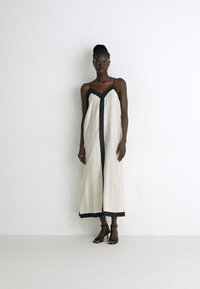 Cremefarbenes ärmelloses Kleid mit schwarzem V-Ausschnitt, seitlichen Schlitzen und schwarzen Saumdetails, aus einem leichten Stoff gefertigt.