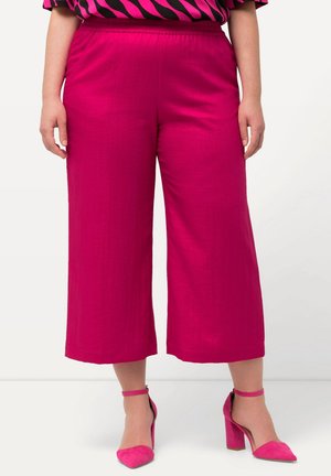 Mujer lleva pantalones anchos de pierna ancha y largos hasta el tobillo en un rosa brillante, con tacones de bloque con correa al tobillo a juego en rosa, combinados con una blusa negra con estampado rosa.