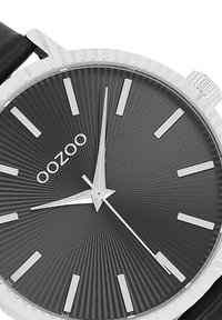 Montre argentée avec un cadran noir texturé, des index et des aiguilles argentés. Elle est dotée d'un bracelet en cuir noir et d'une lunette striée.