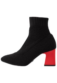 Botín negro de tela con cuello acanalado, punta afilada y un tacón bloque rojo en contraste. Material texturizado con acentos suaves.