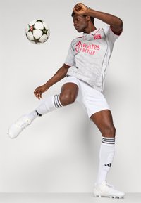 Joueur de football masculin en maillot blanc de Lyon et chaussures à crampons frappant un ballon de football noir et blanc à motif étoilé sur fond uni.