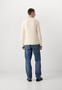 Cream-colored cable-knit sweater with a round neckline, paired with blue denim jeans and black shoes, viewed from the back.