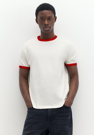 Junger Mann steht mit den Händen in den Taschen, trägt ein weißes T-Shirt mit rotem Kragen und Ärmelsäumen vor einem einfarbigen weißen Hintergrund.