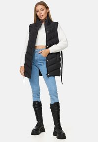 Mustat puffatake, joissa on korkea kaulus, vetoketjukiinnitys ja sivutaskut, päällä valkoinen lyhyt neuletakki ja yhdistettynä vaaleansinihin farkkuihin sekä mustiin saappaisiin.