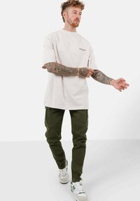 Lichtbeige oversized t-shirt met korte mouwen en een klein merklogo, gecombineerd met donkergroene cargo-broeken en witte sneakers.