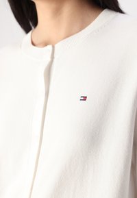 Weißer Knopf-Cardigan aus weichem Stoff, mit einem gerippten Kragen und einem kleinen gestickten Logo in Rot, Weiß und Blau.