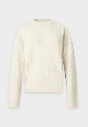 Person, die einen cremefarbenen Strickpullover mit Blattmustern, dunkle weite Hosen und schwarze Schuhe trägt, steht mit einer Hand in der Tasche.