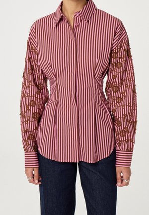 Femme portant une chemise à rayures verticales rose et marron avec une broderie florale marron sur les manches et un jean bleu foncé.