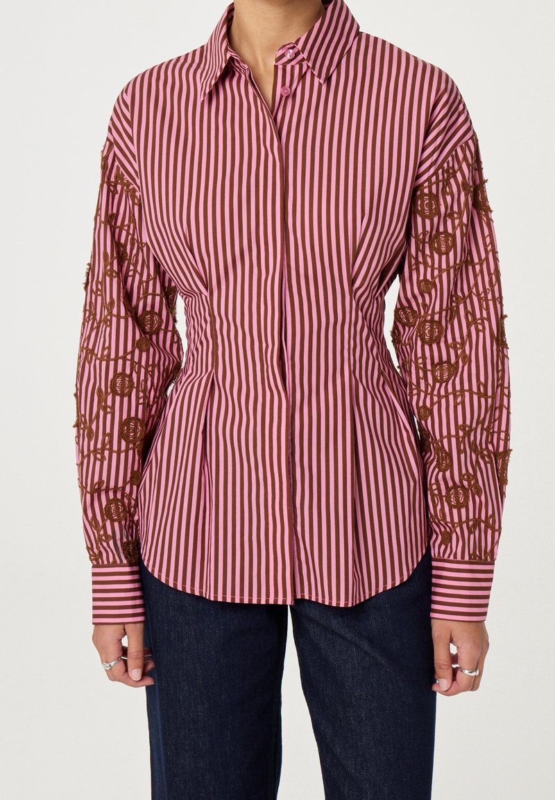 Femme portant une chemise à rayures verticales rose et marron avec une broderie florale marron sur les manches et un jean bleu foncé.