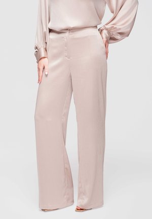 Pantalon large en satin beige porté avec un chemisier assorti à manches longues, les mains reposant sur les hanches contre un fond uni.