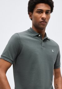 Mladý muž s krátkymi kučeravými vlasmi, oblečený v tmavozelenej krátkorukávovej polokošeli s malým bielym logom na hrudi.