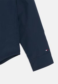Marineblå jakkeærme med en glatt tekstur; har et lite logo i rødt, hvitt og blått ved mansjetten; ren sømdetalj.