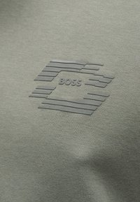 Πράσινο ύφασμα με ανάγλυφο, τμηματικό εξάγωνο σχέδιο και τη λέξη "BOSS" τοποθετημένη στο κέντρο του σχήματος.