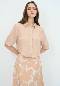 Kurzärmlige, hellrosa Bluse mit Knopfleiste und strukturierter Oberfläche, mit Kragen und abgerundetem Saum. Getragen mit einem fließenden gemusterten Rock.