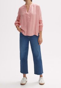 Blouse rose à manches longues avec un col henley et des poignets smockés, associée à un jean bleu court et des baskets blanches.