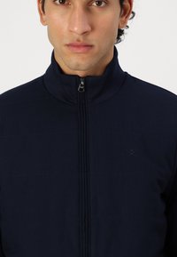 Laivastonsininen vetoketjullinen takki, jossa on korkea kaulus, hienovarainen tekstuurikuvio ja etuvetoketju. Pieni logo kauluksessa.