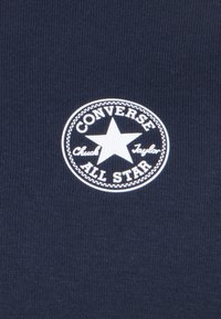 Navy blue fabric with a circular white patch displaying "Converse All Star" and a star in the center, surrounded by decorative stitching.