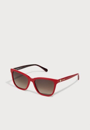 Red sunglasses with oversized square frames, gradient brown lenses, and black patterned arms featuring gold trim and logo detail.