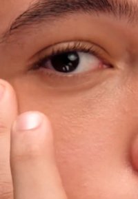 Gros plan d'un œil humain avec une iris foncé et de longs cils. La peau autour de l'œil est claire avec une texture lisse. Un doigt touche doucement la peau à proximité.