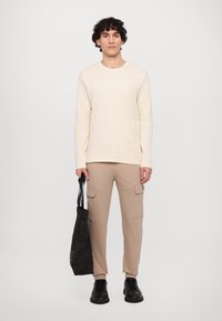 Beige langermet skjorte med strukturert logo, kombinert med beige cargo bukser. Svart veske med korte håndtak i den ene hånden, svarte sko.