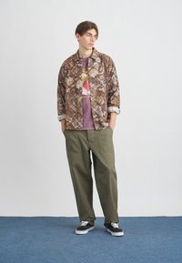 Barna terepmintás ingdzseki lila grafikás pólóval, olívazöld bő szárú nadrággal és fekete tornacipővel párosítva. Fehér bélésű belső ujjak.